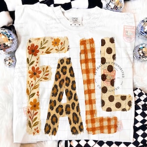 May include: A white short-sleeve t-shirt featuring the word FALL in large letters. The F is decorated with orange flowers and green leaves, the A has a brown leopard print, the L is orange plaid, and the second L has brown polka dots.