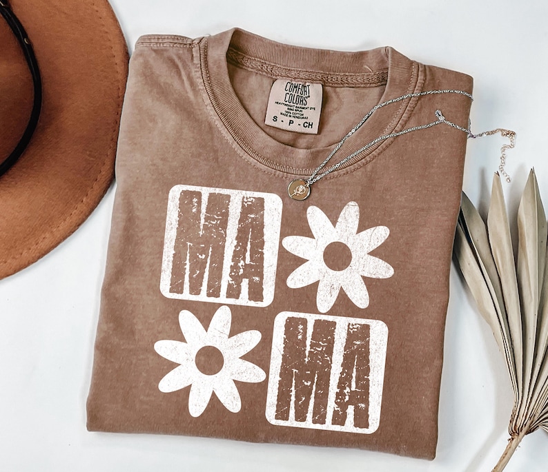Peut inclure: Un t-shirt marron avec un motif blanc qui dit "MAMA" avec des fleurs entre les lettres. Le t-shirt a un aspect vieilli.
