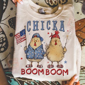 May include: A cream-colored sweatshirt featuring a graphic of two cartoon chickens. One chicken holds an American flag, and the other holds a sparkler. The text "CHICKA BOOM BOOM" is printed across the top and bottom.