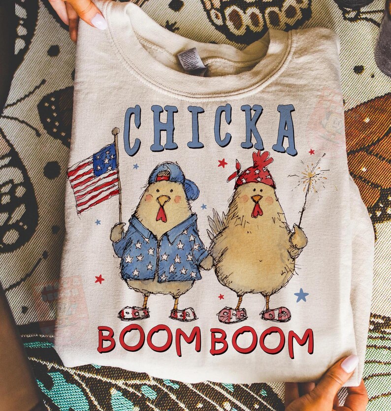 May include: A cream-colored sweatshirt featuring a graphic of two cartoon chickens. One chicken holds an American flag, and the other holds a sparkler. The text "CHICKA BOOM BOOM" is printed across the top and bottom.