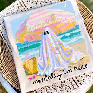 May include: A white t-shirt featuring a graphic of a ghost under a pink and yellow striped beach umbrella on a sandy shore with a straw tote bag and pink heart-shaped sunglasses. The text "mentally i'm here" is printed in black cursive script.