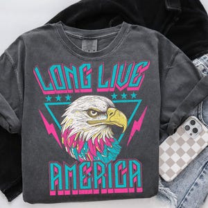 May include: A charcoal grey t-shirt with a graphic print of a bald eagle with pink and blue accents. The text "Long Live America" is printed in pink and blue letters.