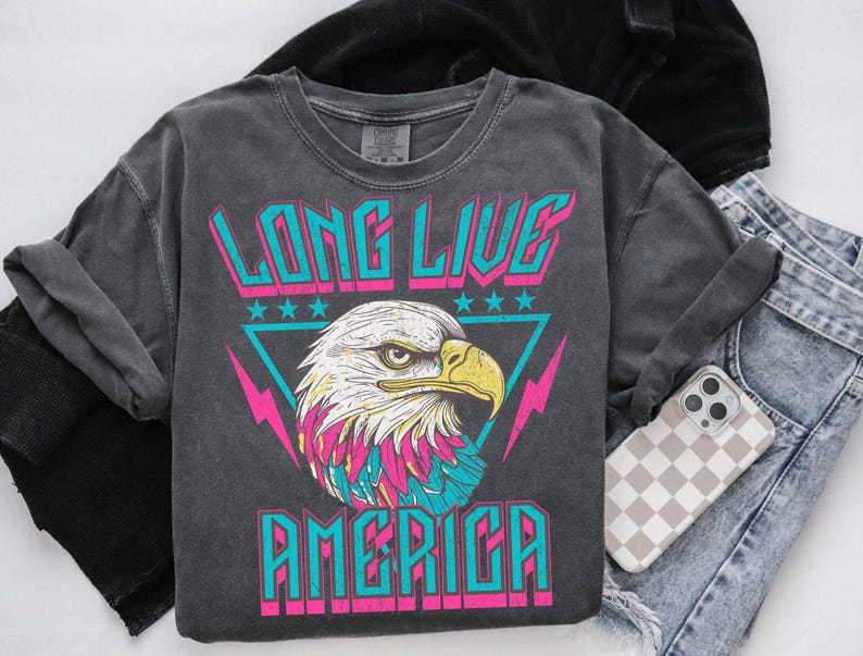 May include: A charcoal grey t-shirt with a graphic print of a bald eagle with pink and blue accents. The text "Long Live America" is printed in pink and blue letters.