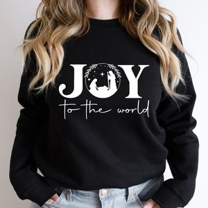 Pu&ograve; includere: Felpa nera con testo bianco che recita "JOY to the world" con un'immagine stilizzata della scena della Nativit&agrave; all'interno della lettera "O".
