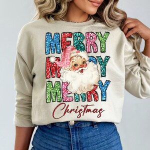Op de afbeelding: Een beige sweatshirt met een feestelijk ontwerp met de woorden "Merry Christmas" in kleurrijke glitterletters en een vintage illustratie van de kerstman met een roze muts.