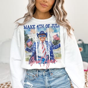 May include: White sweatshirt with a graphic print of a man wearing a cowboy hat and sunglasses, holding American flags. The text "MAKE 4TH OF JULY GREAT AGAIN" is printed on the sweatshirt.