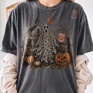 May include: A charcoal gray t-shirt featuring a graphic print of a ghost with a leopard print pattern, a rustic church, a crescent moon, and pumpkins. The design includes a small rectangular badge with the text WHAT A DESIGN.