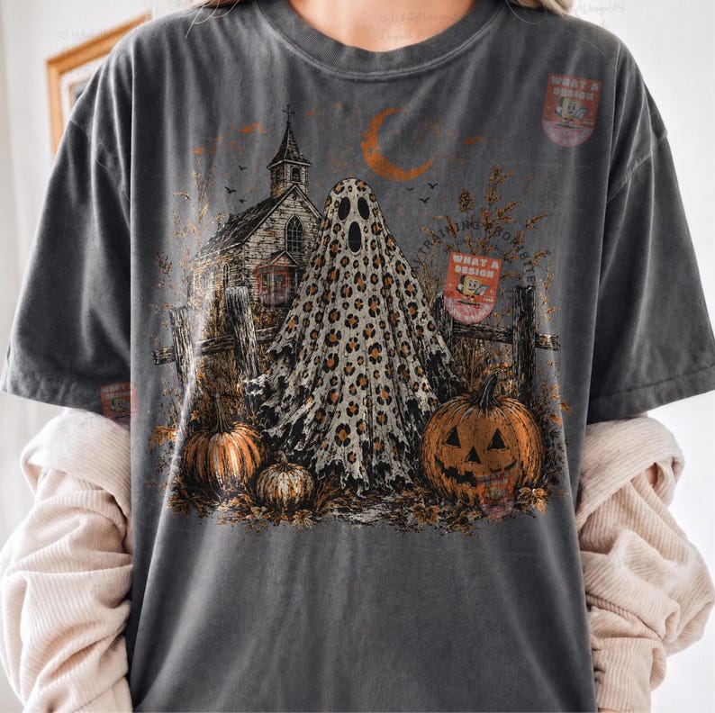 May include: A charcoal gray t-shirt featuring a graphic print of a ghost with a leopard print pattern, a rustic church, a crescent moon, and pumpkins. The design includes a small rectangular badge with the text WHAT A DESIGN.