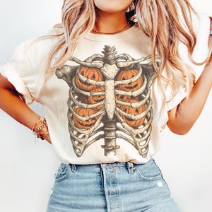 May include: Cream-colored t-shirt featuring a Halloween-themed design. The graphic depicts a skeletal rib cage with orange jack-o'-lanterns inside. The design is detailed and vintage-inspired, perfect for the spooky season.