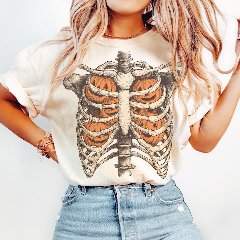 May include: Cream-colored t-shirt featuring a Halloween-themed design. The graphic depicts a skeletal rib cage with orange jack-o'-lanterns inside. The design is detailed and vintage-inspired, perfect for the spooky season.