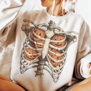 May include: Cream-colored sweatshirt featuring a skeletal rib cage design with orange jack-o'-lanterns inside. The graphic is detailed with bone-colored ribs and orange pumpkins, perfect for Halloween.