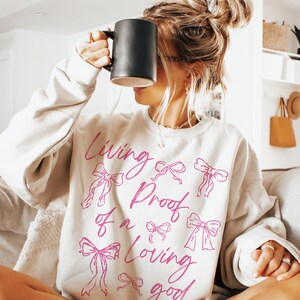 May include: A white sweatshirt with the text "Living Proof of a Loving Good" in pink script. The text is surrounded by pink bows.