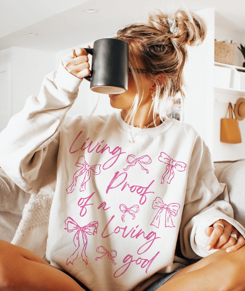May include: A white sweatshirt with the text "Living Proof of a Loving Good" in pink script. The text is surrounded by pink bows.