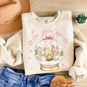 K&ouml;nnte beinhalten: Ein cremefarbener Pullover mit einem rosa-wei&szlig;en Schneekugel-Design. Die Schneekugel zeigt eine Weihnachtskrippe mit Maria, Josef und dem Jesuskind. Der Text "Oh Come, Let us Adore Him" ist um die Schneekugel herum geschrieben.