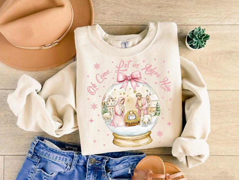 K&ouml;nnte beinhalten: Ein cremefarbener Pullover mit einem rosa-wei&szlig;en Schneekugel-Design. Die Schneekugel zeigt eine Weihnachtskrippe mit Maria, Josef und dem Jesuskind. Der Text "Oh Come, Let us Adore Him" ist um die Schneekugel herum geschrieben.
