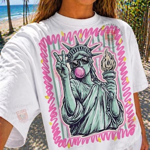 Puede incluir: Camiseta blanca con un gr&aacute;fico de la Estatua de la Libertad con gafas de sol y soplando una burbuja de chicle. El dise&ntilde;o est&aacute; enmarcado en rosa y verde, con detalles dorados. La Estatua de la Libertad sostiene una antorcha y hace la se&ntilde;al de la paz.