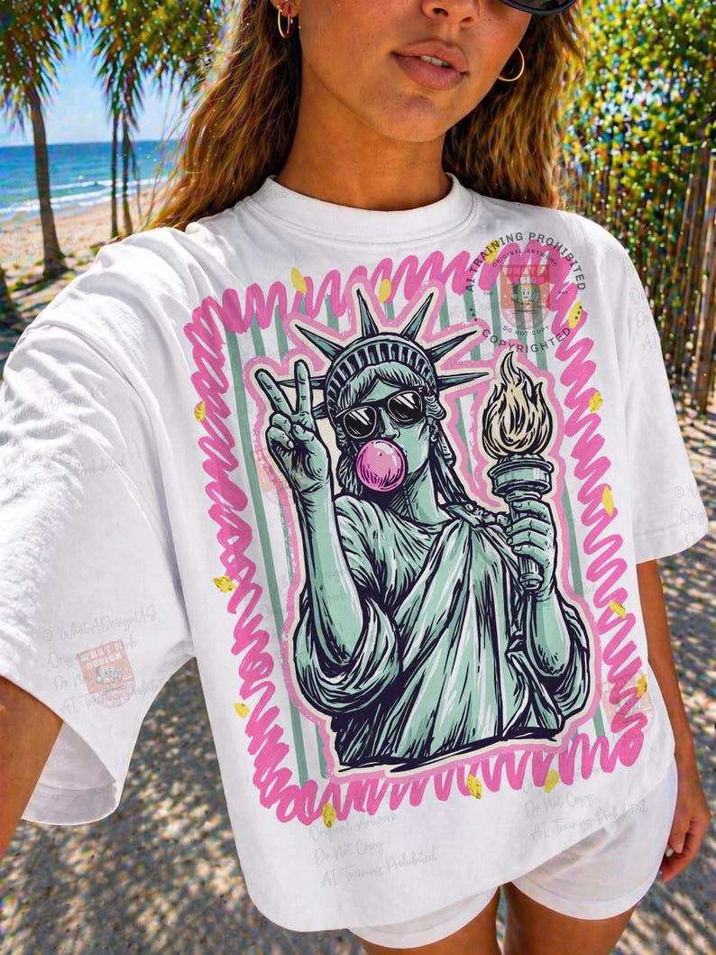 Puede incluir: Camiseta blanca con un gr&aacute;fico de la Estatua de la Libertad con gafas de sol y soplando una burbuja de chicle. El dise&ntilde;o est&aacute; enmarcado en rosa y verde, con detalles dorados. La Estatua de la Libertad sostiene una antorcha y hace la se&ntilde;al de la paz.