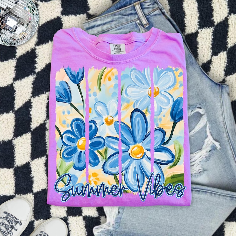 May include: A purple t-shirt with a graphic of blue and white flowers with yellow centers. The flowers are painted with thick brushstrokes and are set against a yellow background. The text "Summer Vibes" is printed in blue at the bottom of the shirt.