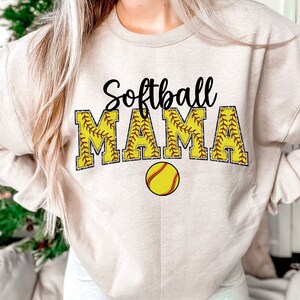 May include: A beige sweatshirt with the text "Softball Mama" in black and yellow. The text is surrounded by a yellow softball with red stitching.