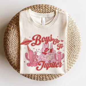 K&ouml;nnte beinhalten: Ein wei&szlig;es T-Shirt mit einem Retro-Grafikdesign, das Cowboys auf Pferden und ein UFO zeigt. Der Text auf dem Shirt lautet "Boys Go To Jupiter".