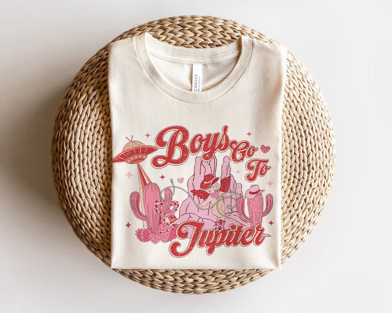 K&ouml;nnte beinhalten: Ein wei&szlig;es T-Shirt mit einem Retro-Grafikdesign, das Cowboys auf Pferden und ein UFO zeigt. Der Text auf dem Shirt lautet "Boys Go To Jupiter".