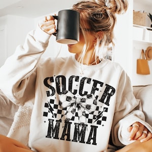 Puede incluir: Una sudadera blanca con un dise&ntilde;o gr&aacute;fico negro que dice "Soccer Mama" con un patr&oacute;n de cuadros blanco y negro y un lazo de bal&oacute;n de f&uacute;tbol.