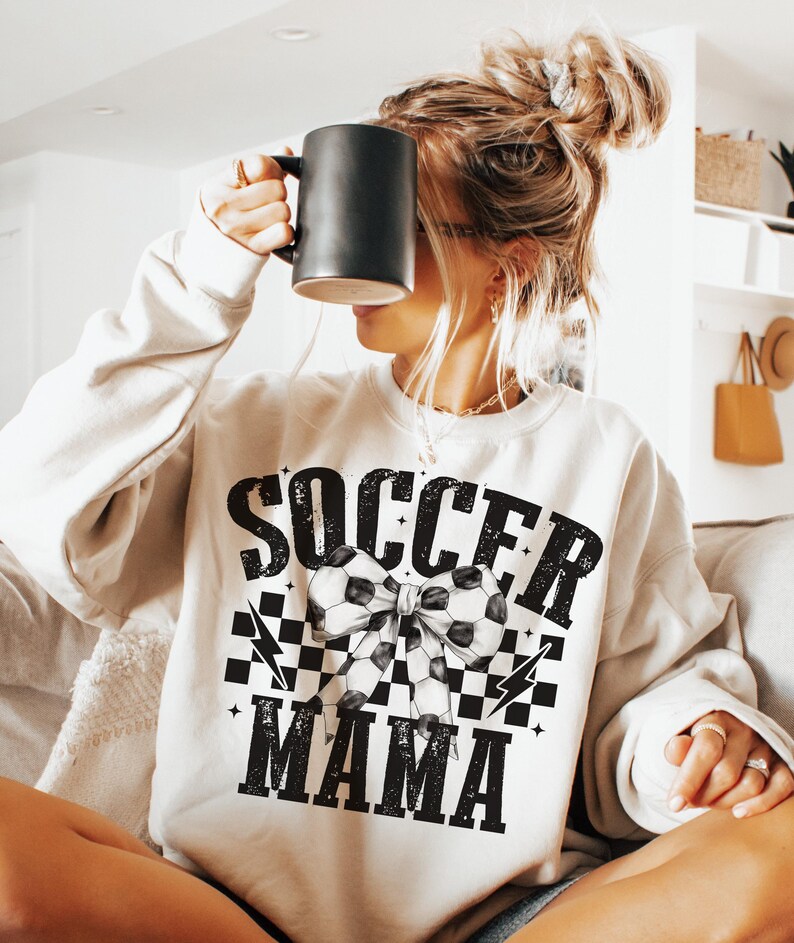 Puede incluir: Una sudadera blanca con un dise&ntilde;o gr&aacute;fico negro que dice "Soccer Mama" con un patr&oacute;n de cuadros blanco y negro y un lazo de bal&oacute;n de f&uacute;tbol.