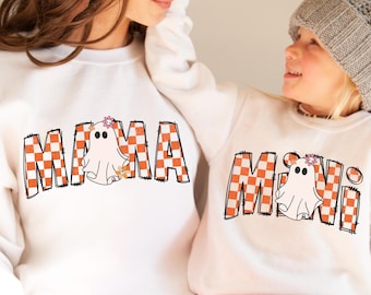 Può includere: Felpe bianche con fantasmi a scacchi arancioni e bianchi. La felpa più grande dice "MAMA" e quella più piccola dice "MINI".