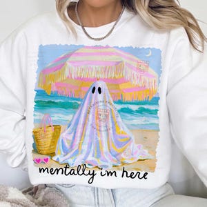 May include: A white crewneck sweatshirt featuring a graphic of a ghost under a pink and yellow striped beach umbrella on a sandy beach with a straw tote bag and pink heart-shaped sunglasses, with the text "mentally i'm here" printed below.