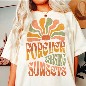 May include: Cream-colored t-shirt featuring a retro-style graphic of a stylized sunburst in shades of orange, peach, and green. The text "FOREVER CHASING SUNSETS" is printed below the sunburst in a matching retro font.