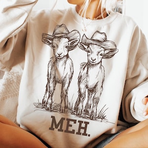 May include: A cream-colored sweatshirt with a graphic of two goats wearing cowboy hats. The goats are drawn in a brown outline and the text "MEH." is printed below them.