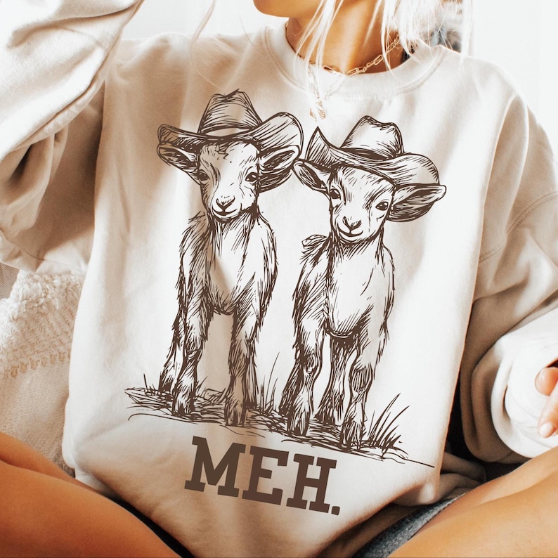 May include: A cream-colored sweatshirt with a graphic of two goats wearing cowboy hats. The goats are drawn in a brown outline and the text "MEH." is printed below them.