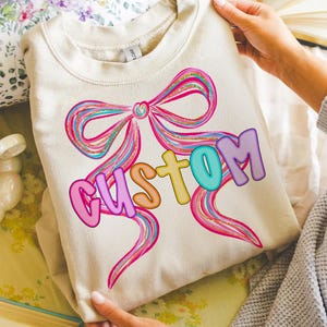 K&ouml;nnte beinhalten: Ein cremefarbenes Sweatshirt mit einem gro&szlig;en, farbenfrohen Schleifendesign mit rosa, blauen und goldenen Akzenten. Das Wort "CUSTOM" ist in Buchstaben auf der Schleife geschrieben. Das Sweatshirt wird von einer Person gehalten.
