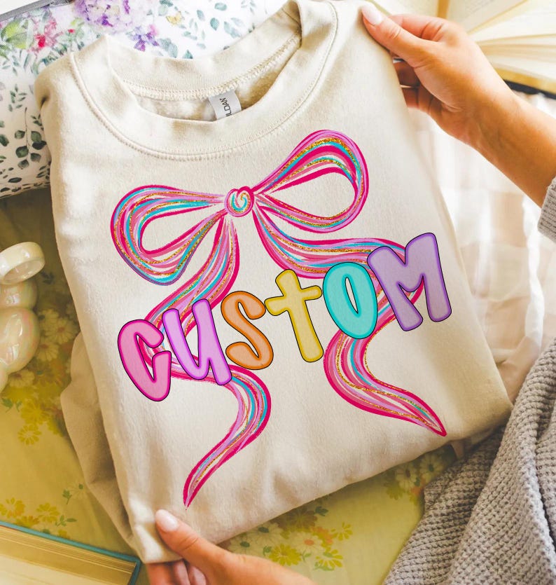 K&ouml;nnte beinhalten: Ein cremefarbenes Sweatshirt mit einem gro&szlig;en, farbenfrohen Schleifendesign mit rosa, blauen und goldenen Akzenten. Das Wort "CUSTOM" ist in Buchstaben auf der Schleife geschrieben. Das Sweatshirt wird von einer Person gehalten.