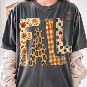 May include: A charcoal gray short-sleeve t-shirt featuring the word FALL printed in large letters. Each letter has a distinct pattern: the F has orange flowers, the A has a leopard print, the L has an orange gingham check, and the L has brown dots.