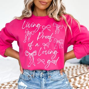 May include: A pink sweatshirt with the text "Living Proof of a Loving Good" in white lettering. The text is surrounded by white bow drawings.