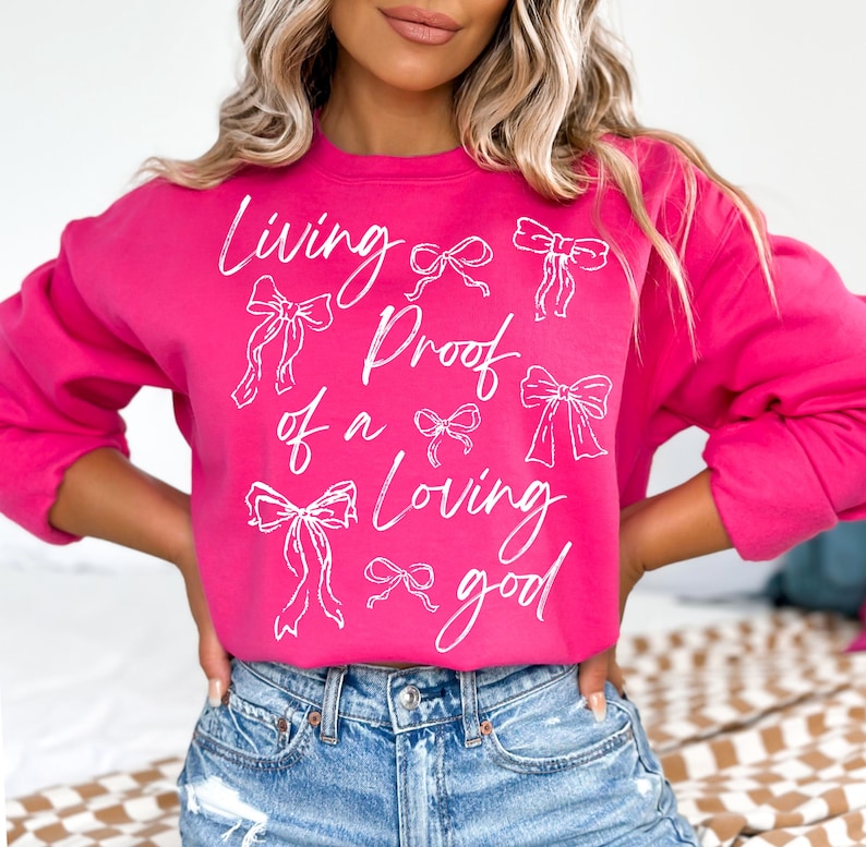 May include: A pink sweatshirt with the text "Living Proof of a Loving Good" in white lettering. The text is surrounded by white bow drawings.