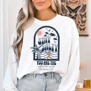 May include: White long-sleeve sweatshirt with a graphic design of a sun, clouds, palm trees, waves, and a person surfing. The text "Stay Salty" is in a retro font. Below the graphic is the text "You are the salt of the earth Matthew 5:13".