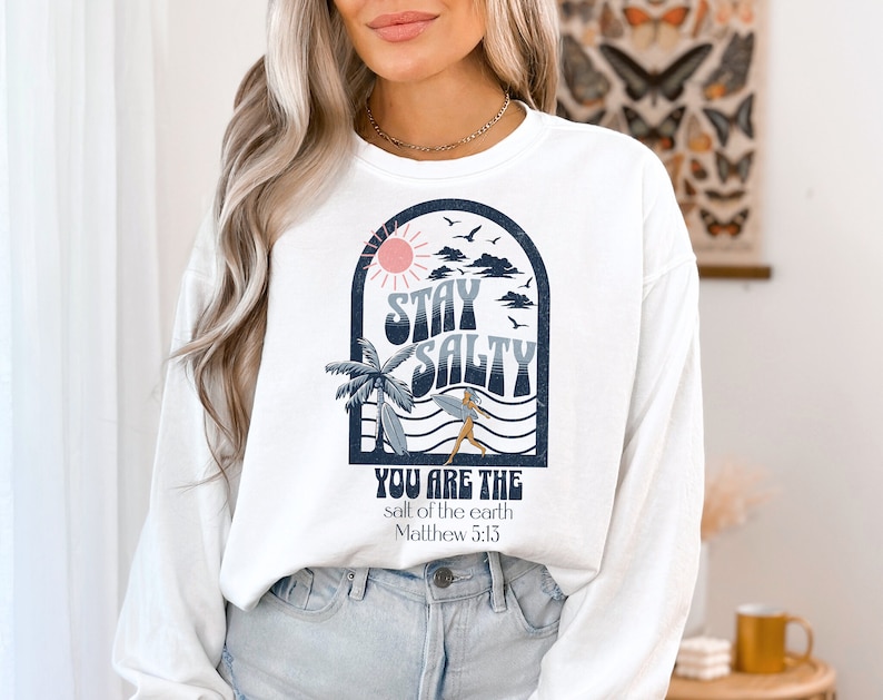 May include: White long-sleeve sweatshirt with a graphic design of a sun, clouds, palm trees, waves, and a person surfing. The text "Stay Salty" is in a retro font. Below the graphic is the text "You are the salt of the earth Matthew 5:13".