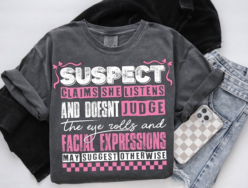 May include: A dark gray t-shirt with a white graphic that says "SUSPECT CLAIMS SHE LISTENS AND DOESNT JUDGE the eye rolls and FACIAL EXPRESSIONS MAY SUGGEST OTHERWISE". The graphic is surrounded by pink ribbon bows.