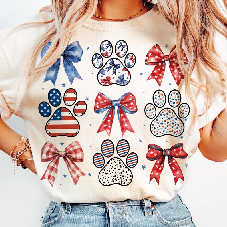 May include: A white t-shirt with a patriotic theme featuring red, white, and blue paw prints and bows. The paw prints are decorated with stars and stripes, while the bows are adorned with red, white, and blue ribbons.