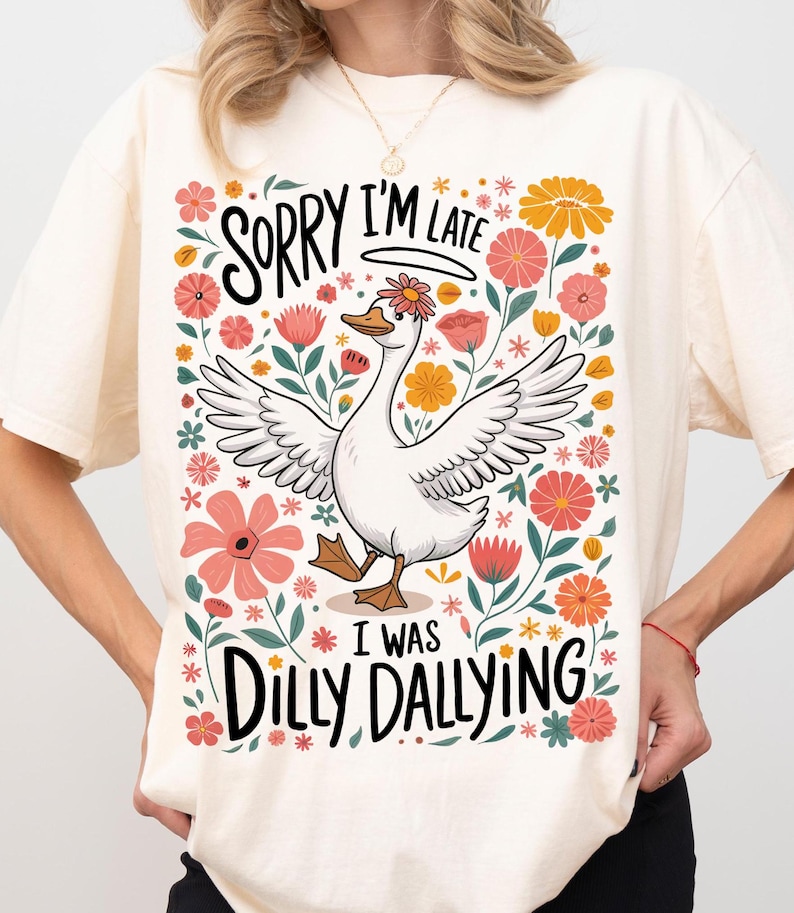 May include: A white t-shirt featuring a floral design and a cartoon goose. The text on the shirt reads "Sorry I'm late I was dilly dallying".