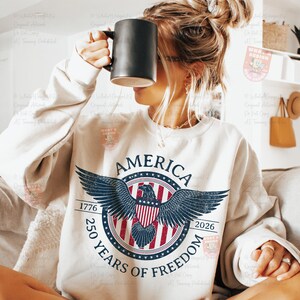 May include: Cream-colored sweatshirt with a patriotic design. The graphic features a circular emblem with an eagle, the text "AMERICA", and "250 YEARS OF FREEDOM" with the years 1776 and 2026. A black mug is held up to the face.