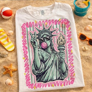 Puede incluir: Camiseta blanca con un gr&aacute;fico de la Estatua de la Libertad con gafas de sol y soplando una burbuja de chicle. El dise&ntilde;o est&aacute; delineado en rosa con un fondo a rayas verdes. La camiseta est&aacute; sobre una superficie arenosa con accesorios de playa.