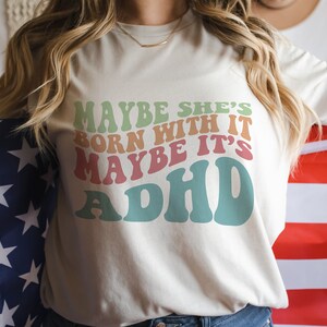 May include: Cream-colored t-shirt with the text "MAYBE SHE'S BORN WITH IT MAYBE IT'S ADHD" in retro-style lettering. The shirt is held in front of an American flag.