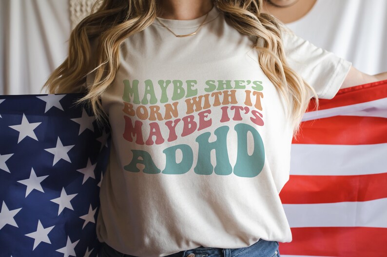 May include: Cream-colored t-shirt with the text "MAYBE SHE'S BORN WITH IT MAYBE IT'S ADHD" in retro-style lettering. The shirt is held in front of an American flag.