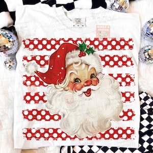 May include: A white t-shirt featuring a graphic of a vintage-style Santa Claus wearing a red hat with white polka dots and a green holly sprig. The background of the graphic consists of horizontal red stripes with white polka dots.