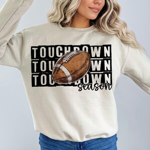 Op de afbeelding: Een beige sweatshirt met een bruine en witte voetbalafbeelding en de tekst "TOUCHDOWN season".