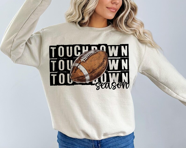 Op de afbeelding: Een beige sweatshirt met een bruine en witte voetbalafbeelding en de tekst "TOUCHDOWN season".