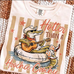 May include: Cream-colored t-shirt with a graphic of an alligator wearing a cowboy hat, sunglasses, and boots, playing a guitar while floating on an inner tube. The text reads "Hotter Than A Hoochie Coochie."